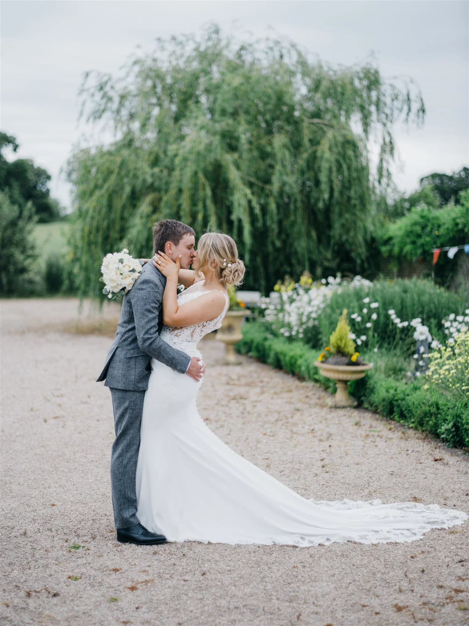 A bride and groom sharing a kiss outdoors on their wedding day, with a large tree and flowers in the background.